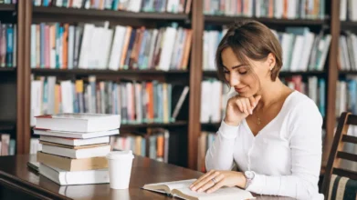 Mulher sentada em mesa de biblioteca lendo um livro, com pilha de livros ao lado e estantes repletas de livros ao fundo. Foto: senivpetro/Freepik