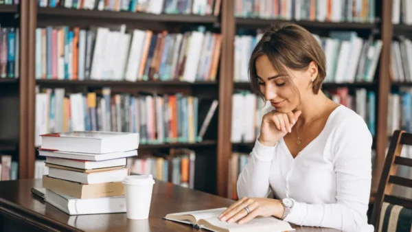 Mulher sentada em mesa de biblioteca lendo um livro, com pilha de livros ao lado e estantes repletas de livros ao fundo. Foto: senivpetro/Freepik