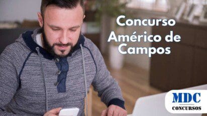 Homem de barba, vestindo moletom cinza com capuz azul, sentado à mesa com um laptop prateado aberto, segurando um smartphone branco e olhando para a tela. O ambiente é uma sala bem iluminada, com móveis de madeira e plantas ao fundo - Américo de Campos (SP) oferece salários de até R$ 2.877 em Concurso Público