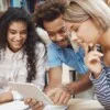 Três pessoas jovens sorridentes analisando juntas um tablet em ambiente de estudo com livros ao fundo. Foto: cookie_studio/Freepik