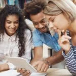 Três pessoas jovens sorridentes analisando juntas um tablet em ambiente de estudo com livros ao fundo. Foto: cookie_studio/Freepik