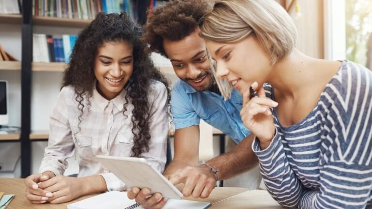 Três pessoas jovens sorridentes analisando juntas um tablet em ambiente de estudo com livros ao fundo. Foto: cookie_studio/Freepik