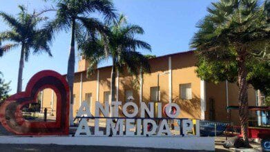 Fotografia tirada durante o dia mostra um letreiro turístico com a frase "Antônio Almeida PI", posicionado em frente a um prédio de fachada amarela com janelas brancas e telhado vermelho. Ao lado esquerdo do letreiro há um coração estilizado em vermelho. O local é arborizado, com palmeiras altas e outras árvores, sugerindo um ambiente urbano e acolhedor no município de Antônio Almeida, no estado do Piauí.