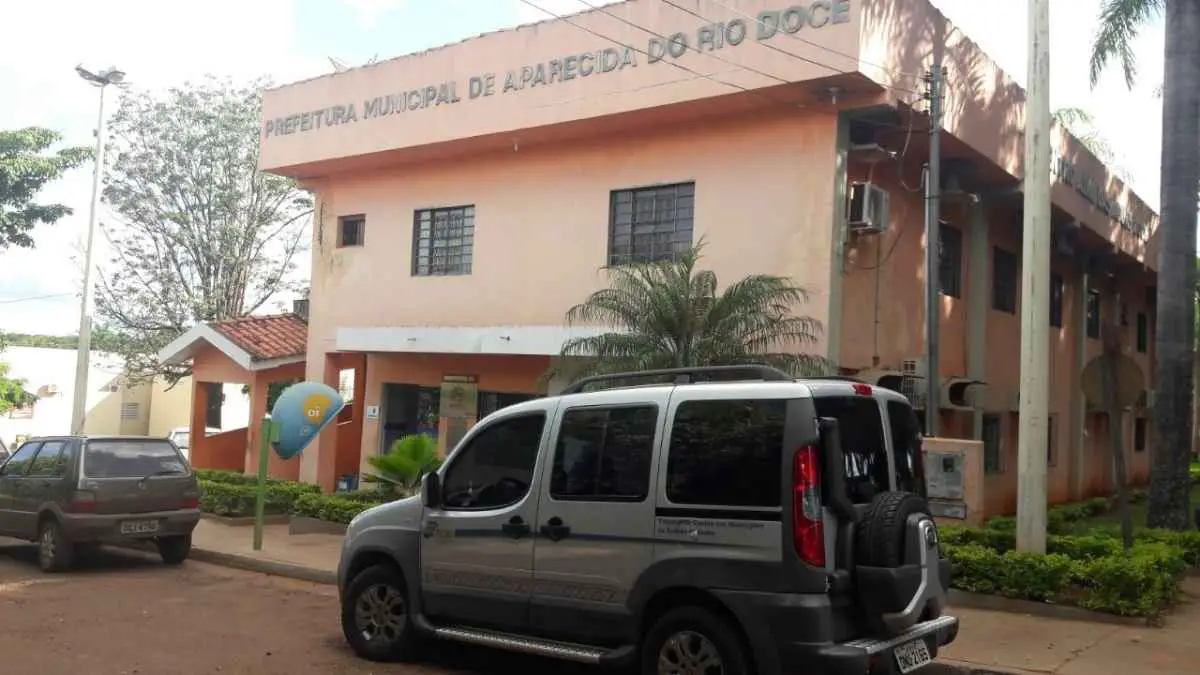 Fachada da Prefeitura Municipal de Aparecida do Rio Doce, GO, com veículos estacionados em frente e uma cabine telefônica da Oi na calçada.