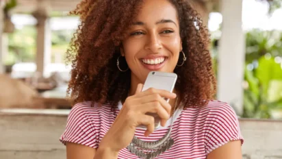 Mulher sorridente de cabelos cacheados segura um celular enquanto está sentada em ambiente externo. Foto: wayhomestudio/Freepik