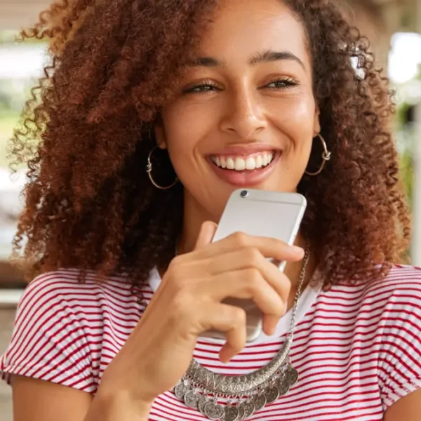 Mulher sorridente de cabelos cacheados segura um celular enquanto está sentada em ambiente externo. Foto: wayhomestudio/Freepik