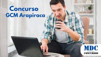 Homem de pele clara e cabelo curto escuro, usando camisa xadrez azul e branca sobre camiseta cinza, sentado em uma poltrona rosa. Ele segura um copo de café na mão direita e usa um notebook preto sobre uma mesa branca. O ambiente é iluminado, com decoração minimalista e estante branca ao fundo - Arapiraca abre 48 vagas para Guarda Municipal