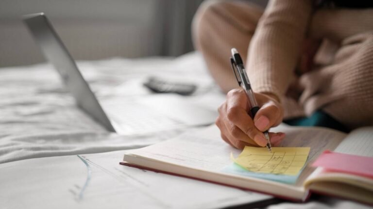 Pessoa sentada em uma cama usando roupas confortáveis escreve em um post-it amarelo enquanto estuda com livros, cadernos e gráficos abertos ao redor. Na frente dela, há um notebook parcialmente visível e uma calculadora, sugerindo um ambiente de estudo ou trabalho remoto. A imagem transmite foco, organização e dedicação, com destaque para a mão segurando uma caneta e anotando informações. (Araraquara)