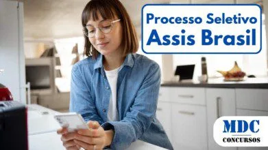 Mulher jovem com óculos e cabelo castanho liso, vestindo camisa jeans e camiseta branca, segurando um smartphone e olhando para a tela com expressão tranquila. Ela está em uma cozinha moderna e iluminada, com bancada, frutas e um laptop ao fundo - Assis Brasil/AC abre vagas em Processo Seletivo (Foto: Freepik)
