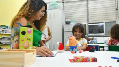 Professora acompanha criança sorridente durante atividade lúdica com brinquedos educativos em sala de aula infantil, com materiais pedagógicos sobre a mesa e outros alunos ao fundo. Foto: Divulgação/Prefeitura de Bacabeira-MA