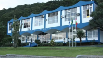 Prédio da Prefeitura Municipal de Balneário Camboriú com fachada azul e branca, bandeiras hasteadas e área arborizada ao redor. Foto: Divulgação/Prefeitura de Balneário Camboriú-SC