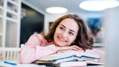 Imagem de uma jovem sorridente com cabelos castanhos ondulados e blusa rosa clara, apoiando o queixo nas mãos sobre um livro aberto em uma mesa. Ela parece estar em um ambiente de estudo moderno, com estantes ao fundo e iluminação suave no teto. O clima é descontraído e positivo, transmitindo motivação, concentração e leveza no momento de leitura ou estudo.