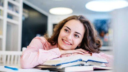 Imagem de uma jovem sorridente com cabelos castanhos ondulados e blusa rosa clara, apoiando o queixo nas mãos sobre um livro aberto em uma mesa. Ela parece estar em um ambiente de estudo moderno, com estantes ao fundo e iluminação suave no teto. O clima é descontraído e positivo, transmitindo motivação, concentração e leveza no momento de leitura ou estudo.