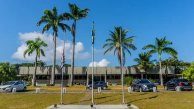 Fachada da sede da Prefeitura de Bertioga com palmeiras, três mastros com bandeiras e carros estacionados em frente sob céu azul. Foto: Divulgação/Prefeitura de Bertioga-SP
