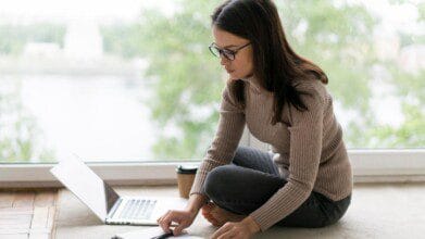 Mulher sentada no chão usando laptop e anotações, com copo de café ao lado, em ambiente iluminado por janela ampla. Foto: Freepik