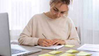 Mulher escrevendo em caderno sobre mesa com laptop, folhas e post-its amarelos. Foto: Freepik