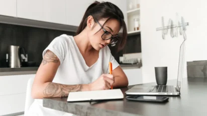 Mulher usando óculos escreve em caderno enquanto estuda com notebook sobre a mesa em ambiente doméstico moderno. Foto: drobotdean/Freepik