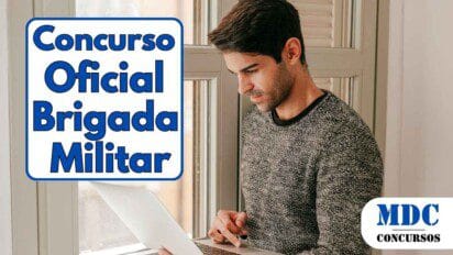 Homem de cabelos escuros usando suéter cinza escuro, em pé perto de uma janela, concentrado enquanto usa um notebook prata - Brigada Militar (RS) lança Concurso com 120 vagas para Oficial