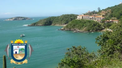 Vista panorâmica de Armação dos Búzios com mar azul-esverdeado, enseada cercada por morros verdes, barcos ancorados e construções à beira-mar, com o brasão oficial do município no canto inferior esquerdo. Foto: Mariordo Mario Roberto Duran Ortiz/Wikipédia