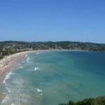 Vista panorâmica de uma praia movimentada em Armação dos Búzios, com mar azul, faixa de areia clara e morros ao fundo.