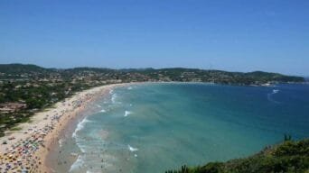 Vista panorâmica de uma praia movimentada em Armação dos Búzios, com mar azul, faixa de areia clara e morros ao fundo.