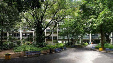 Vista do pátio arborizado do CEFET/RJ, com bancos azuis, jardins e prédio da instituição ao fundo.