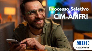 Processos Seletivos 7 Homem sorridente com barba e óculos de armação verde segura um smartphone enquanto se apoia em uma almofada mostarda. Ele veste uma camisa verde sobre uma camiseta branca. O ambiente tem iluminação aconchegante com luzes quentes e frias, móveis modernos e decoração ao fundo - CIM-AMFRI abre vagas em Processo Seletivo