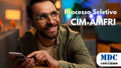 Homem sorridente com barba e óculos de armação verde segura um smartphone enquanto se apoia em uma almofada mostarda. Ele veste uma camisa verde sobre uma camiseta branca. O ambiente tem iluminação aconchegante com luzes quentes e frias, móveis modernos e decoração ao fundo - CIM-AMFRI abre vagas em Processo Seletivo