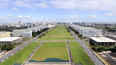 Vista aérea da Esplanada dos Ministérios, em Brasília, com prédios governamentais alinhados e extensas áreas verdes sob céu com poucas nuvens