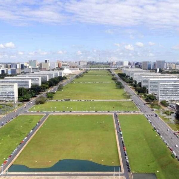 Vista aérea da Esplanada dos Ministérios, em Brasília, com prédios governamentais alinhados e extensas áreas verdes sob céu com poucas nuvens