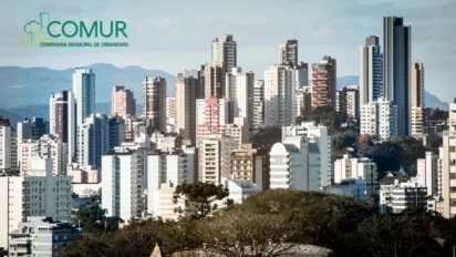 Vista urbana de Novo Hamburgo com prédios residenciais e comerciais ao fundo, vegetação em primeiro plano e logotipo da COMUR no canto superior esquerdo. Foto: Samuel Quintana/CMNH/Wikipédia