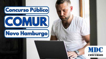 Homem com barba curta e camiseta branca sentado perto de uma janela, concentrado enquanto digita em um notebook prata - COMUR Novo Hamburgo (RS) abre vagas em Concurso Público 2025
