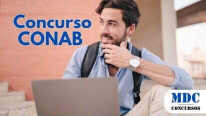 Homem jovem com barba e cabelo escuro sentado ao ar livre com um laptop. Ele veste camisa azul clara, calça bege e mochila preta. Usa um relógio prateado no pulso e segura o queixo com a mão, sorrindo levemente. O fundo mostra uma parede de tijolos e degraus, sugerindo um ambiente universitário ou urbano - CONAB abre 403 vagas com salários de até R$ 8.140 em Concurso