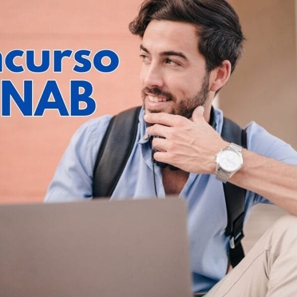 Homem jovem com barba e cabelo escuro sentado ao ar livre com um laptop. Ele veste camisa azul clara, calça bege e mochila preta. Usa um relógio prateado no pulso e segura o queixo com a mão, sorrindo levemente. O fundo mostra uma parede de tijolos e degraus, sugerindo um ambiente universitário ou urbano - CONAB abre 403 vagas com salários de até R$ 8.140 em Concurso