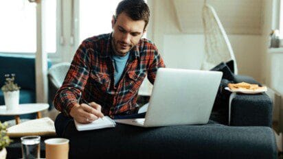 Homem sentado no sofá estudando com caderno e notebook, ao lado de uma mesa com café e pão tostado. Foto: Drazen Zigic/Freepik