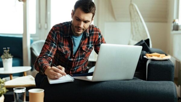 CORE-SE abre concurso público com 131 vagas de até R$ 4 mil 5 Homem sentado no sofá estudando com caderno e notebook, ao lado de uma mesa com café e pão tostado. Foto: Drazen Zigic/Freepik