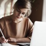 Mulher jovem com óculos escrevendo em um caderno enquanto usa um laptop em uma mesa.