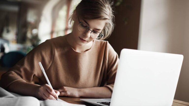 Mulher jovem com óculos escrevendo em um caderno enquanto usa um laptop em uma mesa.