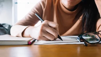 Pessoa escrevendo em um caderno sobre a mesa com óculos e livros ao lado. Foto: Freepik