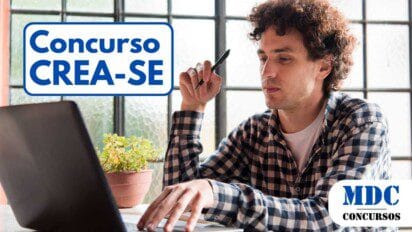 Homem jovem de cabelos cacheados e pele clara, vestindo camisa xadrez, trabalha em um notebook enquanto segura uma caneta na mão. Ele parece concentrado, olhando para a tela. O ambiente tem luz natural, com uma grande janela ao fundo e um vaso com planta sobre a mesa - CREA SE oferece Remuneração de Até R$ 8,8 mil em novo Concurso Público