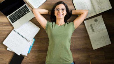 Mulher de óculos deitada no chão sorrindo, cercada por cadernos, livros abertos e um notebook, aparentando estar estudando ou revisando conteúdo.