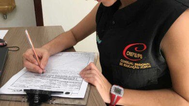 Pessoa preenchendo um formulário sobre uma prancheta, vestindo uniforme do CREF10/PB com o logo do Conselho Regional de Educação Física.