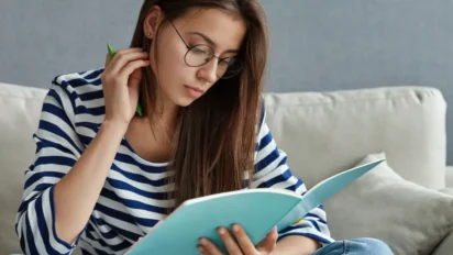Mulher jovem com óculos e blusa listrada estuda em um caderno azul enquanto segura uma caneta verde. Foto: wayhomestudio/Freepik