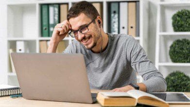 Homem sorridente com óculos e fones de ouvido estuda em frente a um notebook, com livros e cadernos sobre a mesa, em ambiente com prateleiras ao fundo.