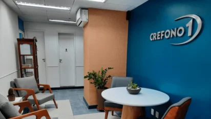 Ambiente interno do CREFONO 1 com área de espera, cadeiras, mesa redonda, plantas decorativas e parede azul com logotipo do conselho. Foto: Divulgação/CREFONO 1