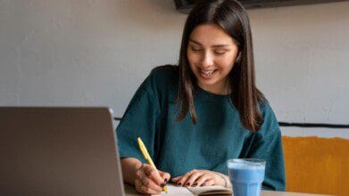 CRM DF abre concurso c/ 270 vagas e salários de até R$ 11 mil 5 Mulher sorridente escrevendo em um caderno, sentada à mesa com um notebook aberto e um copo com bebida azul.