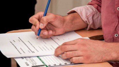 Pessoa usando camisa rosa preenche um formulário de prova ou inscrição com lápis azul em uma carteira escolar. A imagem foca nas mãos e no documento, que contém campos para nome, assinatura e outras informações pessoais. O ambiente sugere uma situação de avaliação, concurso público ou exame oficial. O cenário transmite seriedade e concentração no preenchimento correto dos dados exigidos - CRMV PI