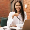 Mulher sorrindo com camisa branca segura óculos enquanto estuda com livros abertos e notebook sobre a mesa em ambiente interno com parede de tijolos. Foto: ArthurHidden/Freepik