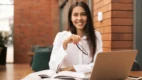 Mulher sorrindo com camisa branca segura óculos enquanto estuda com livros abertos e notebook sobre a mesa em ambiente interno com parede de tijolos. Foto: ArthurHidden/Freepik