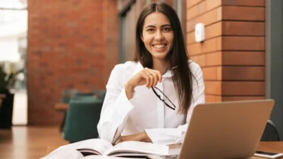 Mulher sorrindo com camisa branca segura óculos enquanto estuda com livros abertos e notebook sobre a mesa em ambiente interno com parede de tijolos. Foto: ArthurHidden/Freepik